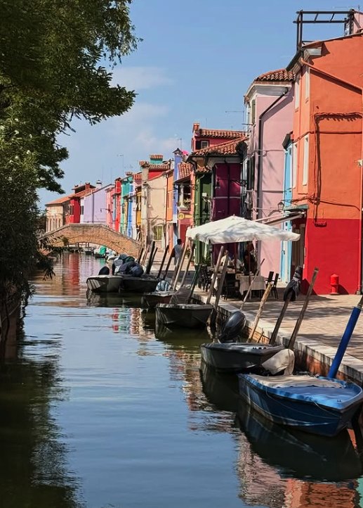A Burano canal curving past a low brick bridge, with a cafe parasol and moored boats along the quay