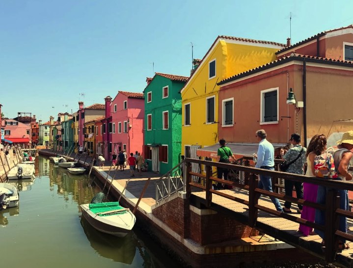 Visitors crossing a wooden bridge over a Burano canal lined with yellow, green and pink houses