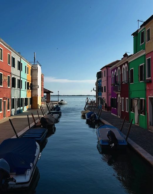 A Burano canal lined with moored boats looking out to the open Venetian Lagoon