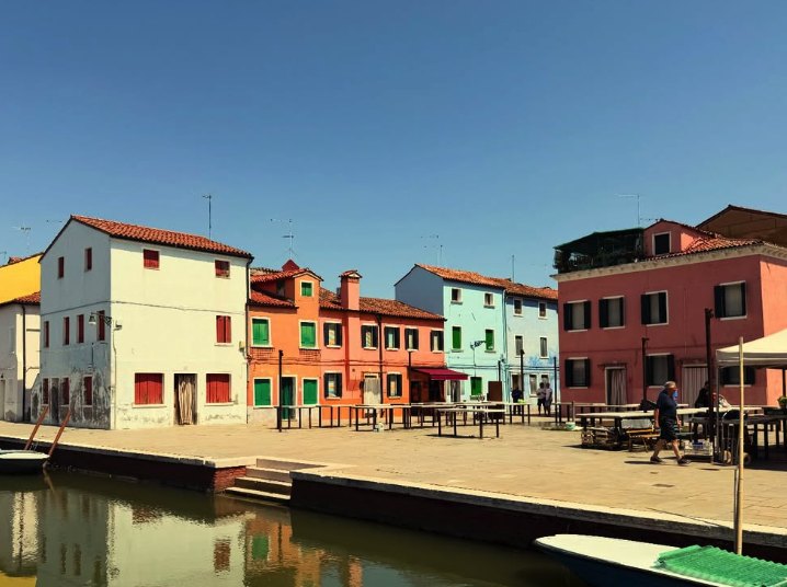 A row of white, orange, mint and pink houses facing a Burano canal beside an open square