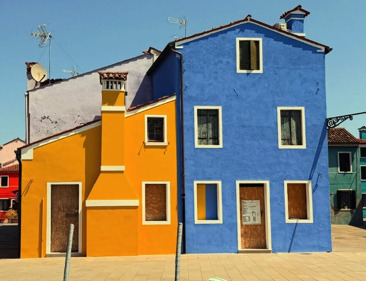 An orange house and a blue house standing side by side on a Burano square under a clear sky