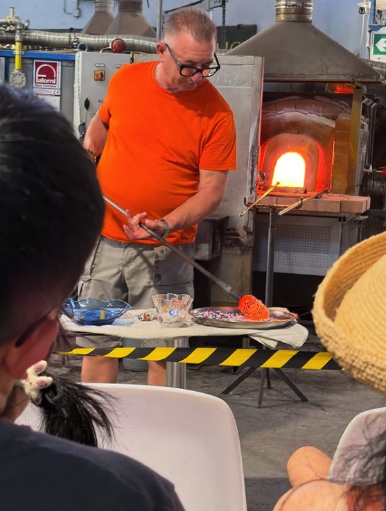 A glassblower shaping a piece at the furnace while a seated audience watches the demonstration